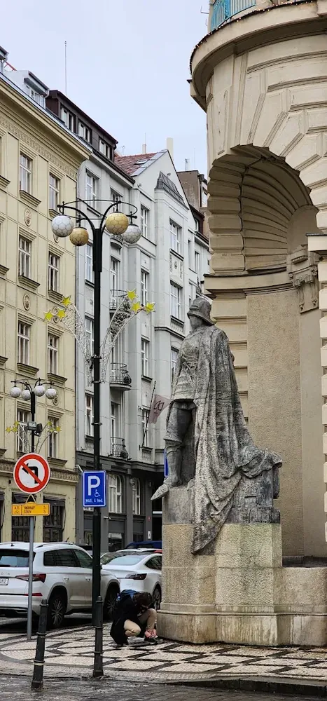 Estatua de un caballero con capa en una calle urbana frente a edificios clásicos y un farola decorada.