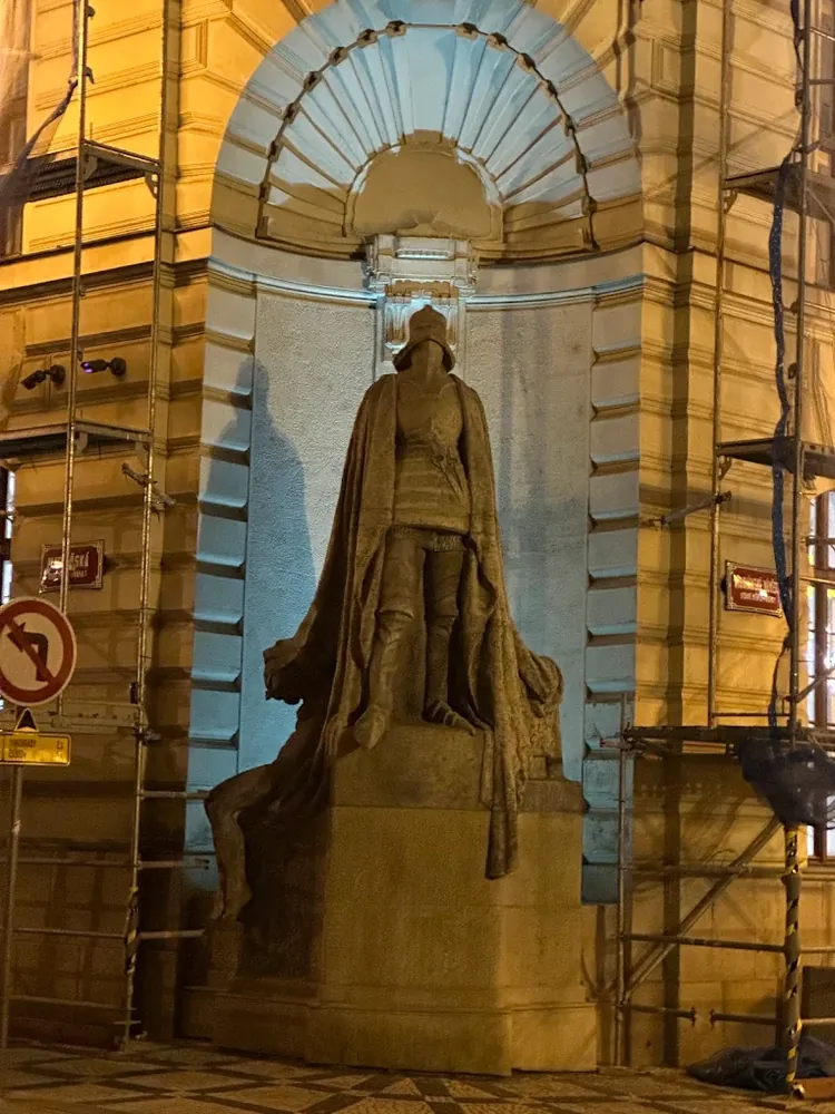 Estatua oscura de un caballero con armadura y capa dentro de un nicho abovedado, flanqueada por andamios a la luz nocturna.