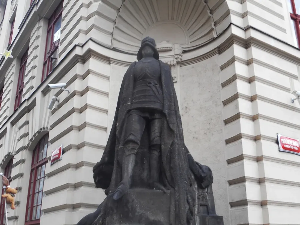Estatua oscura de un caballero con armadura y capa, ubicada en la esquina de un edificio clásico de fachada blanca.