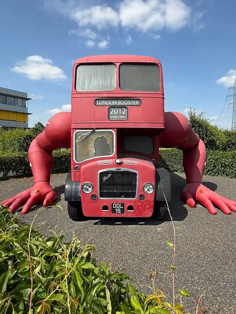 Escultura London Booster roja con grandes brazos articulados apoyados en el suelo, sobre grava y vegetación.