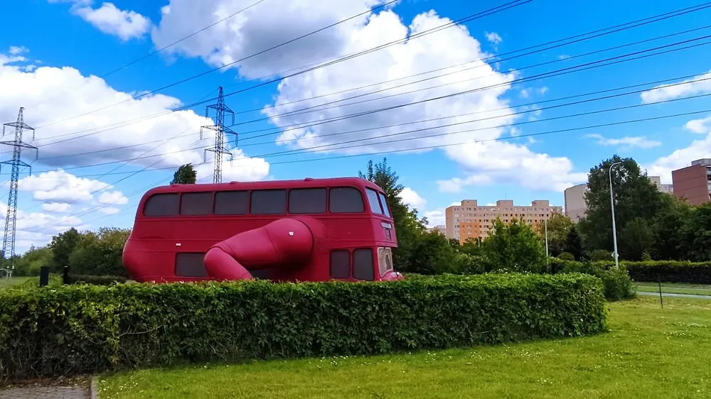 Autobús rojo de dos pisos modificado con un brazo mecánico gigante, sobre un seto verde, bajo un cielo nublado.
