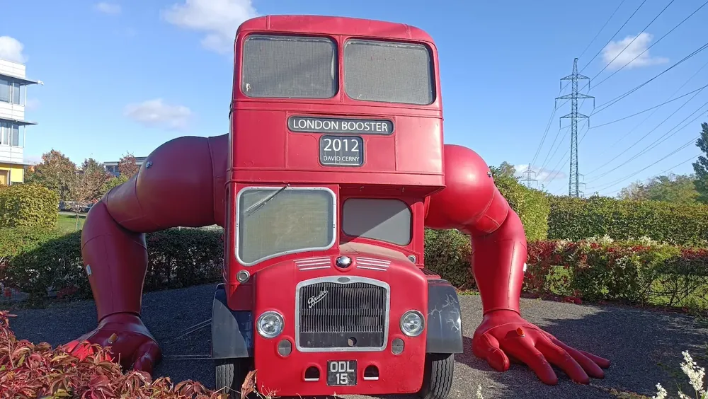 Escultura roja de un autobús londinense de dos pisos con brazos articulados apoyados en el suelo, bajo un cielo azul con n...