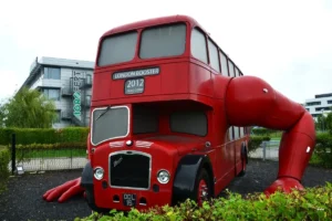 Autobús rojo de dos pisos con brazos musculosos gigantes integrados, expuesto al aire libre junto a vegetación y edificios.