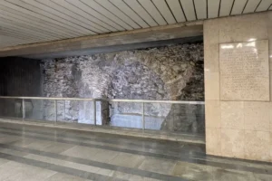 Restos de muros de piedra con arcos protegidos por una barandilla de cristal en el interior de una estación, con una placa...