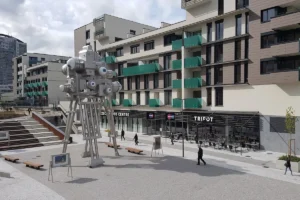 Gran escultura metálica de una cámara de cine con ojos en una plaza frente al edificio TRIFOT y comercios.