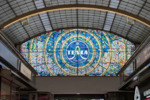 Gran vitral circular en tonos azules y amarillos con el texto "TESLA Radio" en el centro, ubicado bajo un techo abovedado.