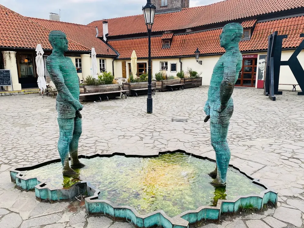 Dos estatuas de hombres en una fuente con forma de mapa sobre un patio empedrado, frente a edificios de tejados rojos.