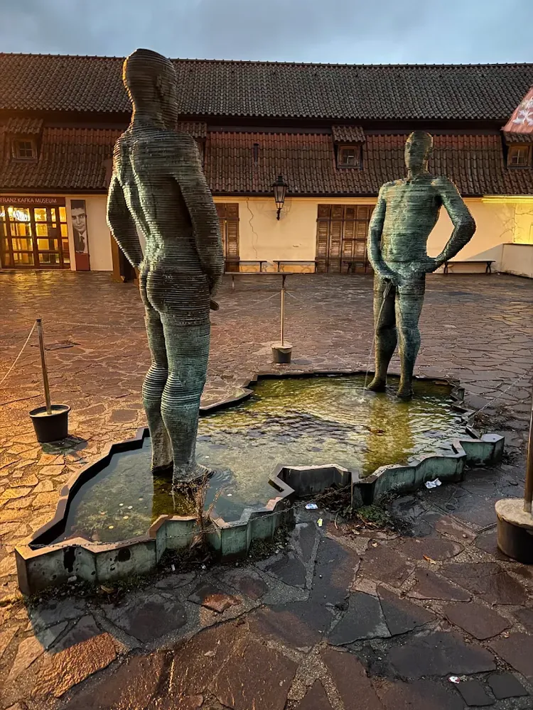 Dos esculturas metálicas de hombres de pie en una fuente con forma de mapa, en un patio empedrado al atardecer.