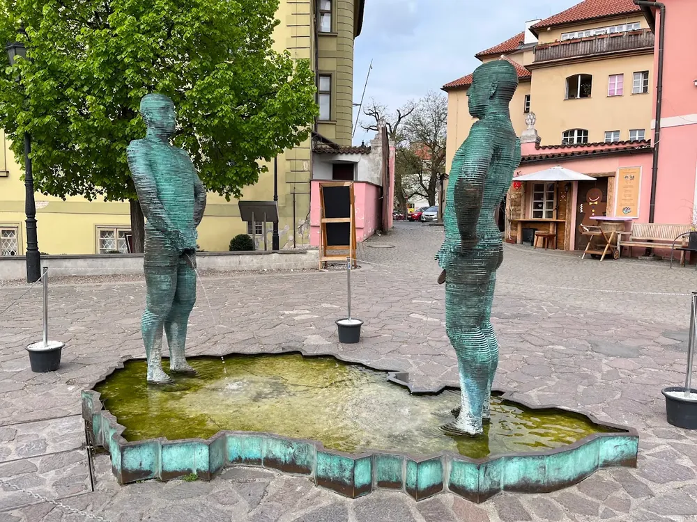 Dos estatuas de bronce en una plaza adoquinada, sobre una fuente con la forma de la República Checa; una de ellas orina.