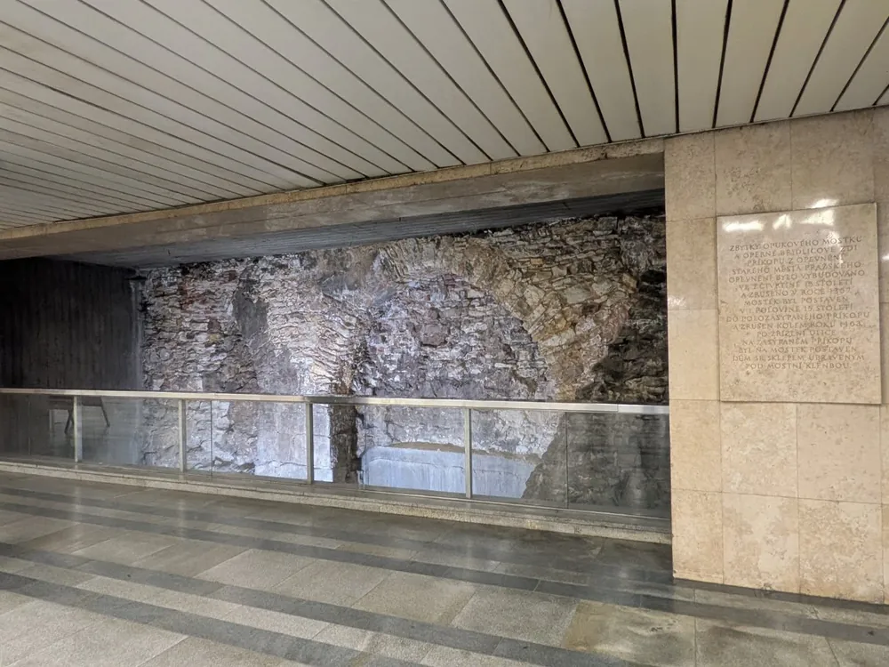Vestigio de puente medieval de piedra protegido por una barandilla de cristal en una estación de metro con una placa conme...