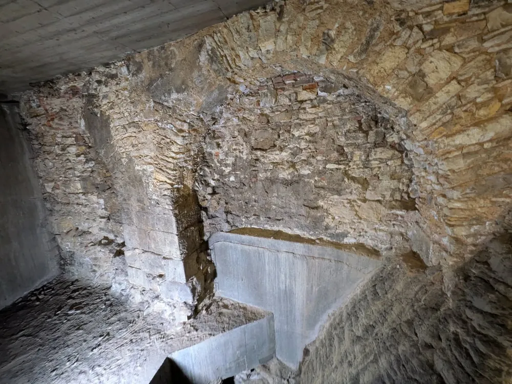 Estructura interior subterránea con muros de piedra irregular, arcos y elementos de hormigón gris.