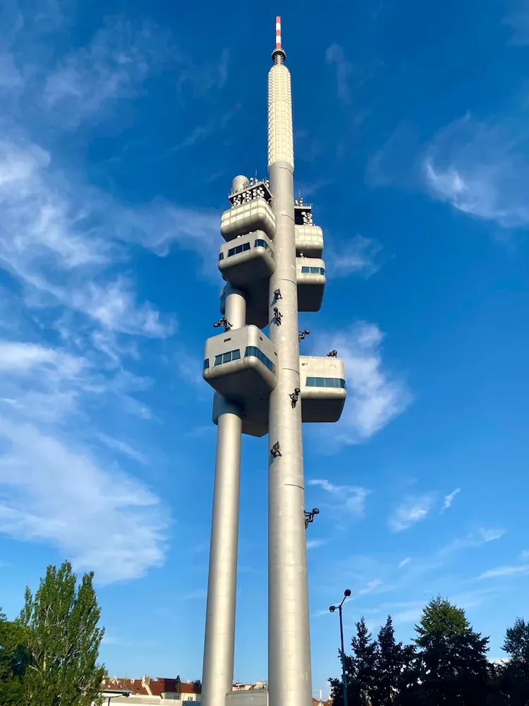 Torre de telecomunicaciones gris con cápsulas y esculturas de bebés trepando, bajo un cielo azul con nubes ligeras.