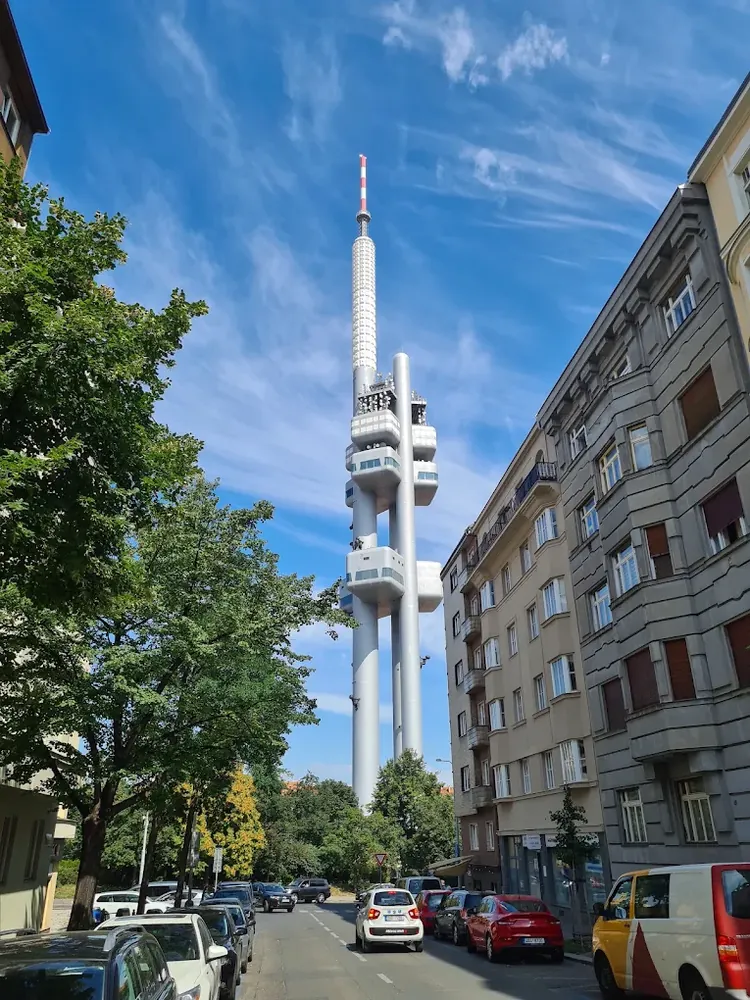 Torre Žižkov vista desde una calle estrecha flanqueada por edificios residenciales y árboles, bajo un cielo azul con nubes...