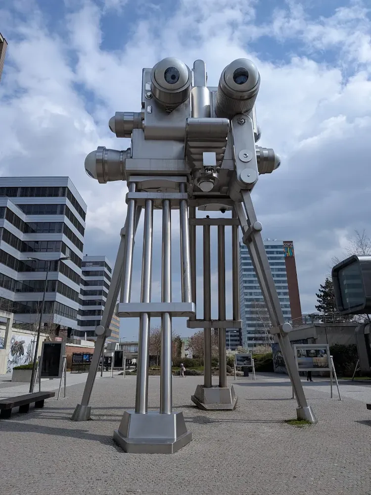 Escultura metálica de una cámara antigua con grandes ojos en un espacio público al aire libre rodeado de edificios.
