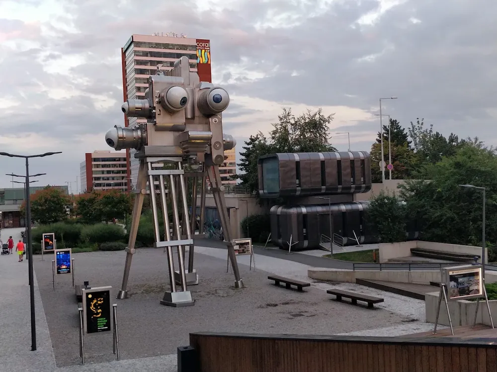 Escultura metálica de una cámara con ojos en un parque urbano, con edificios al fondo bajo un cielo nublado.