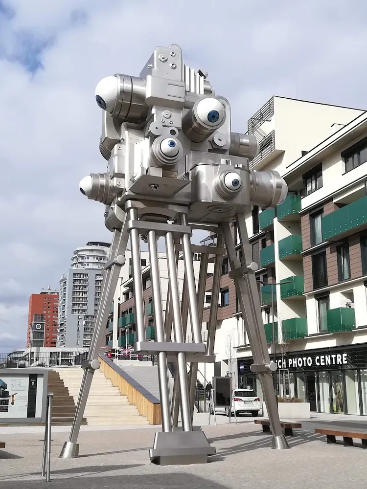 Gran escultura metálica en forma de cámara de cine con múltiples ojos, apoyada sobre un trípode gigante en una plaza urbana.