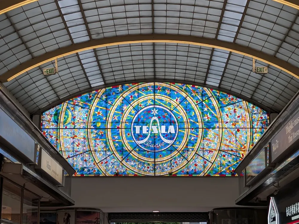 Gran vitral circular en tonos azules y amarillos con el texto "TESLA Radio" en el centro, bajo un techo de cristal abovedado.