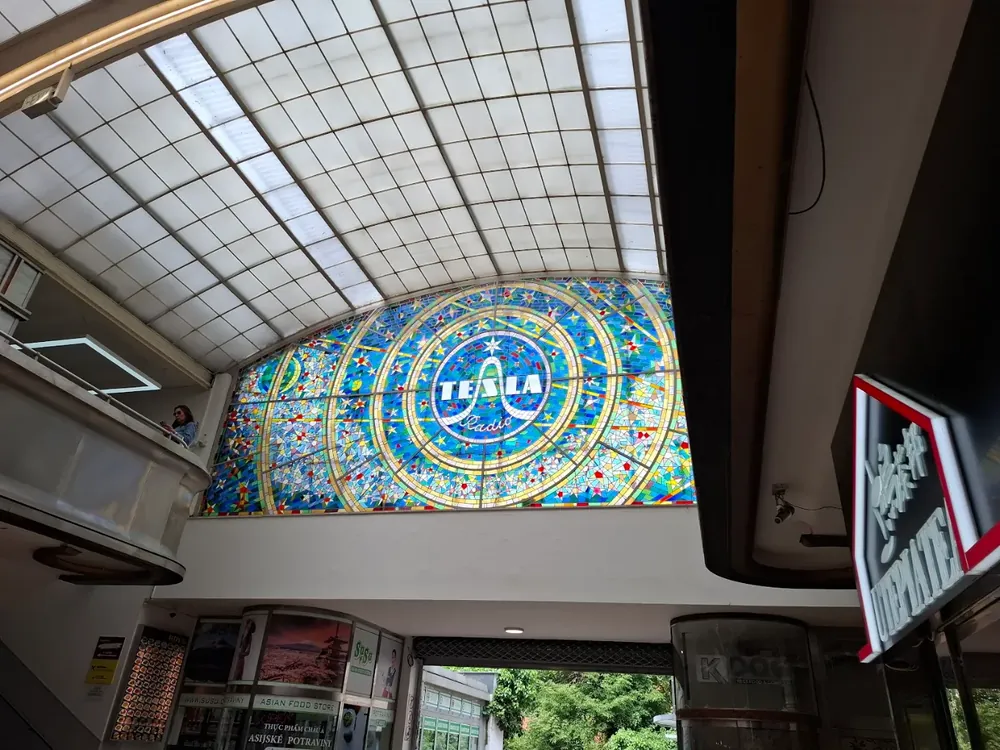 Gran vitral circular azul y amarillo con la inscripción "TESLA Radio" en un interior iluminado con tragaluz en cuadrícula.